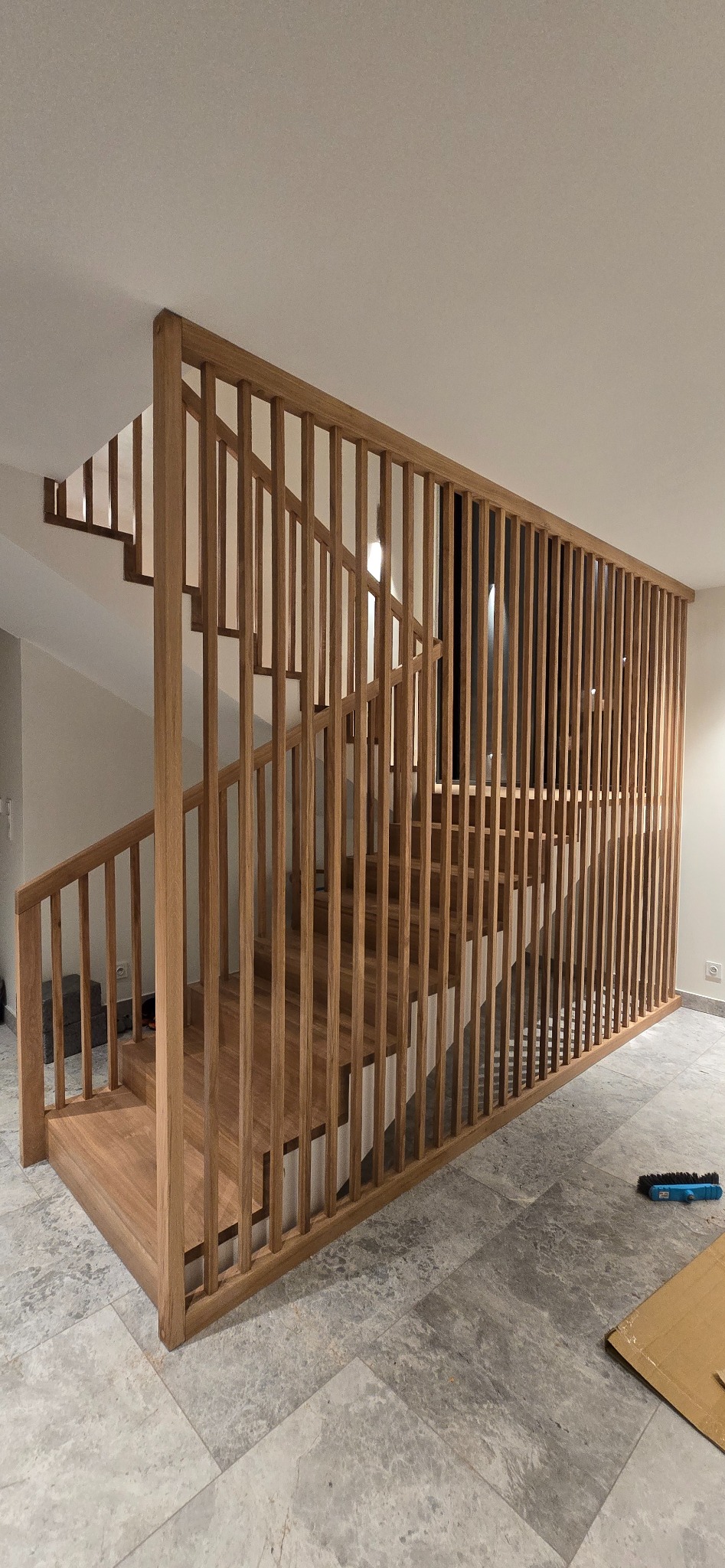 Nowoczesne schody z drewnianą balustradą z pionowych listew, widok z dołu, jasne wnętrze, szare płytki podłogowe, minimalistyczny design.