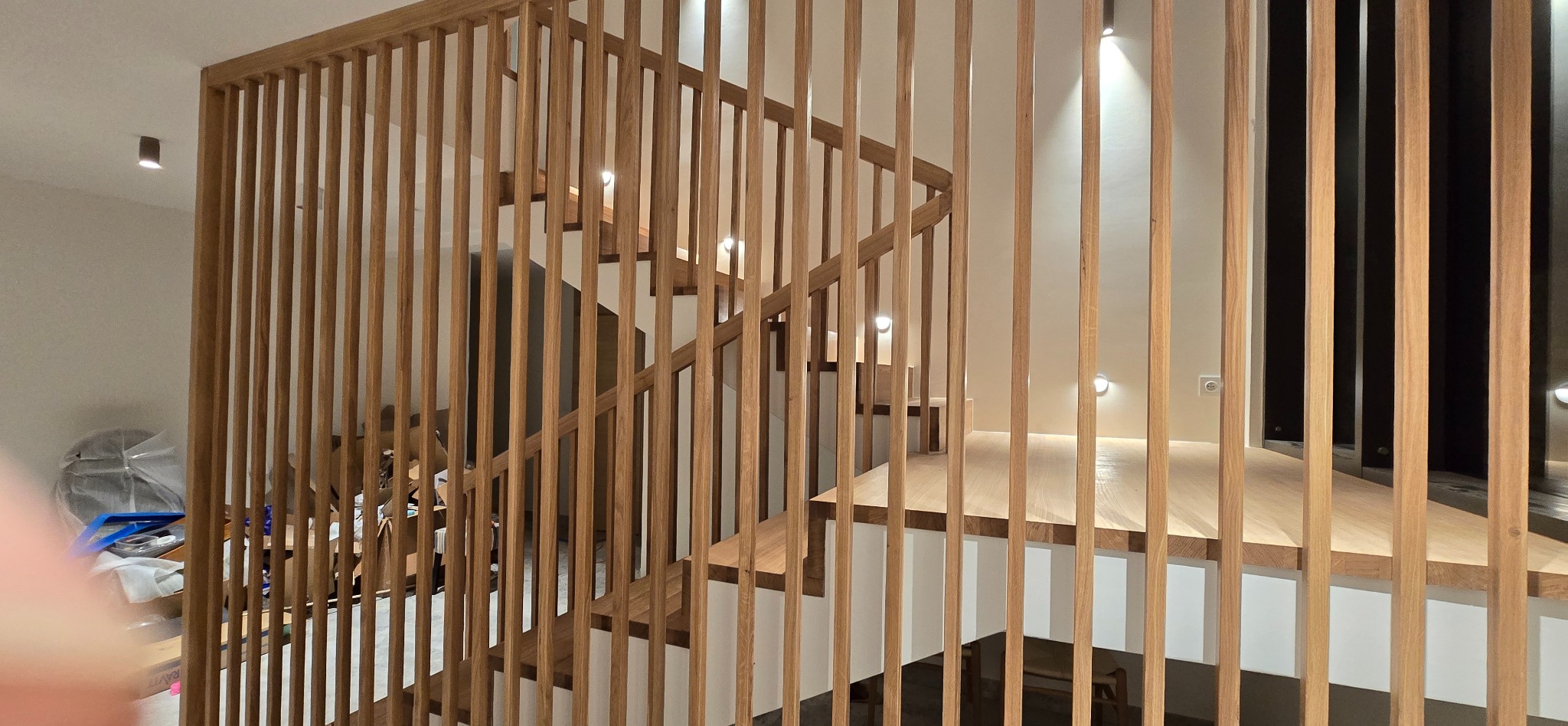 Ażurowe schody z drewna dębowego, z pionowymi listwami tworzącymi balustradę. Widoczne stopnie i oświetlenie punktowe. Minimalistyczny design, ciepłe odcienie drewna.