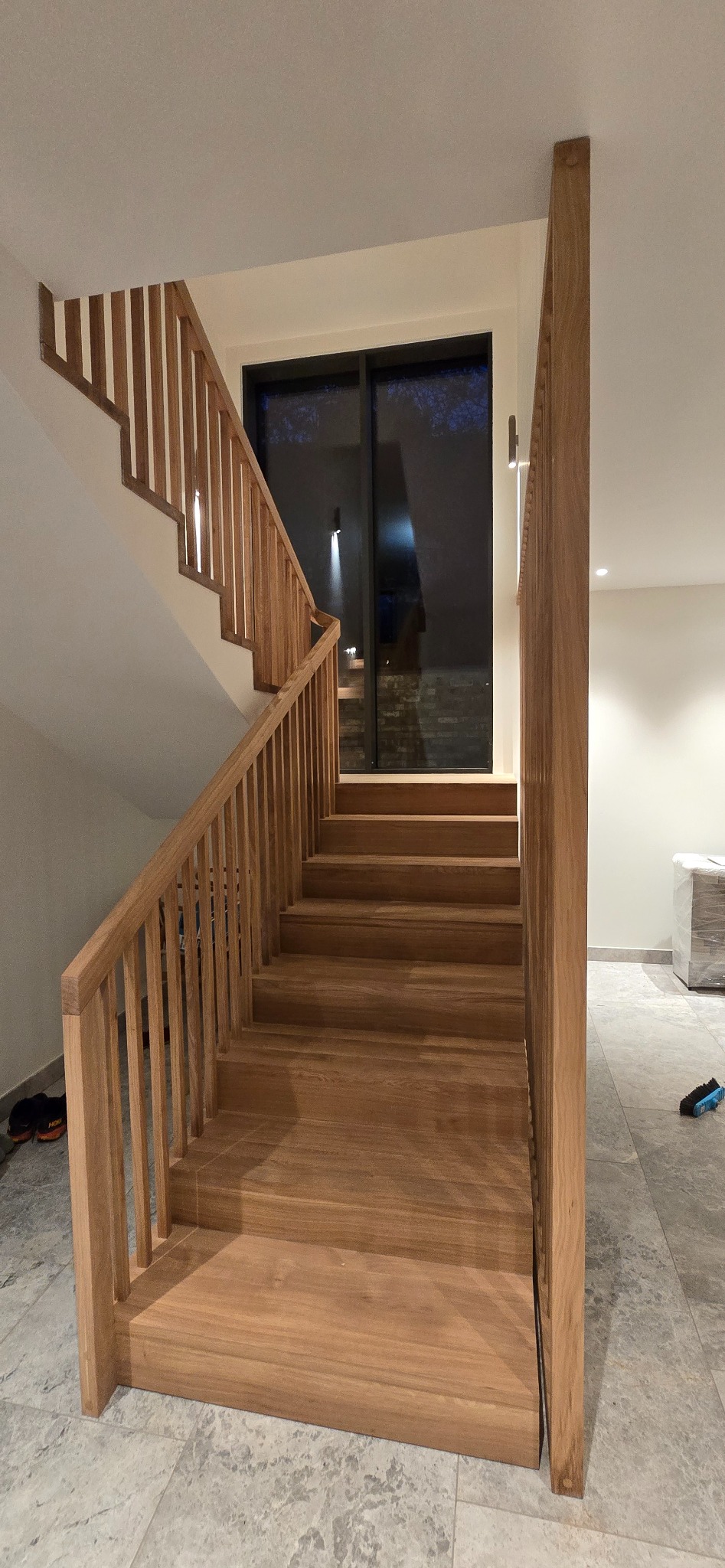 Jasne, drewniane schody z balustradą z pionowych szczebli, prowadzące do okna. Widoczne buty i szczotka na podłodze z szarych płytek.