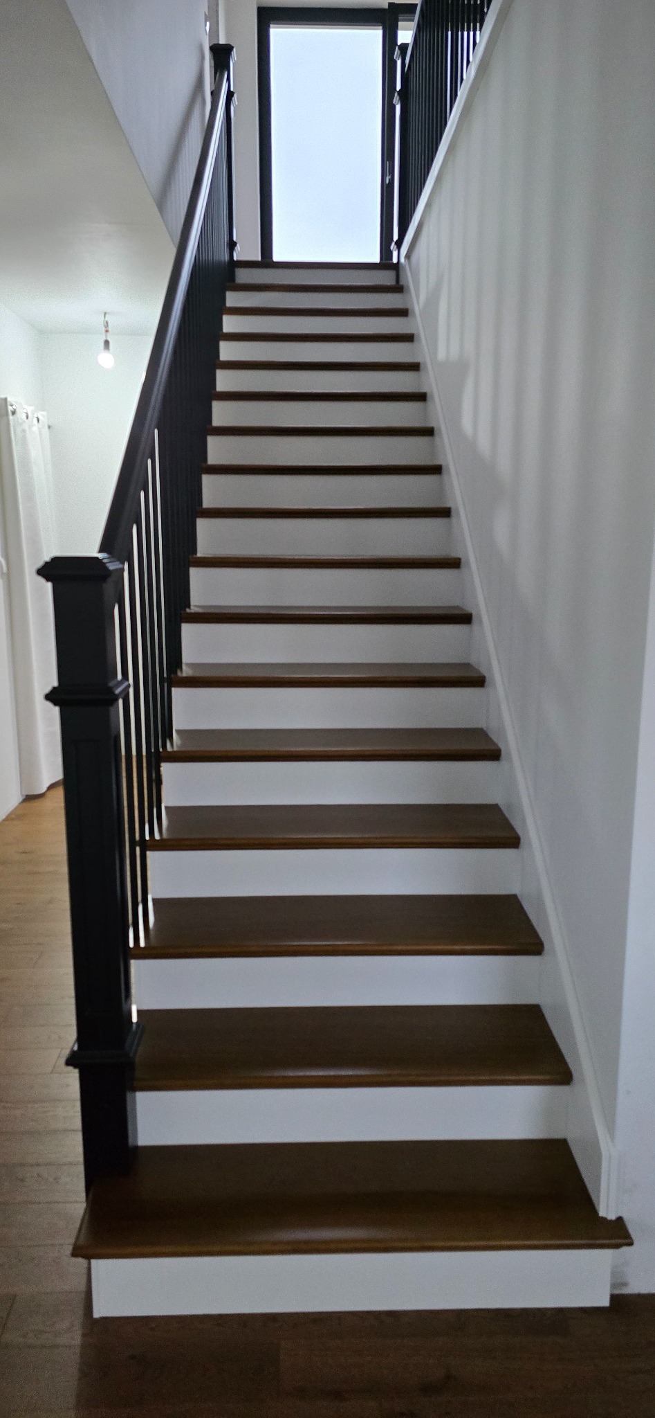 Klasyczne schody z drewnianymi stopniami i białą podstopnicą, czarna balustrada. Widok z dołu na górę, w tle okno z widokiem na zewnątrz.