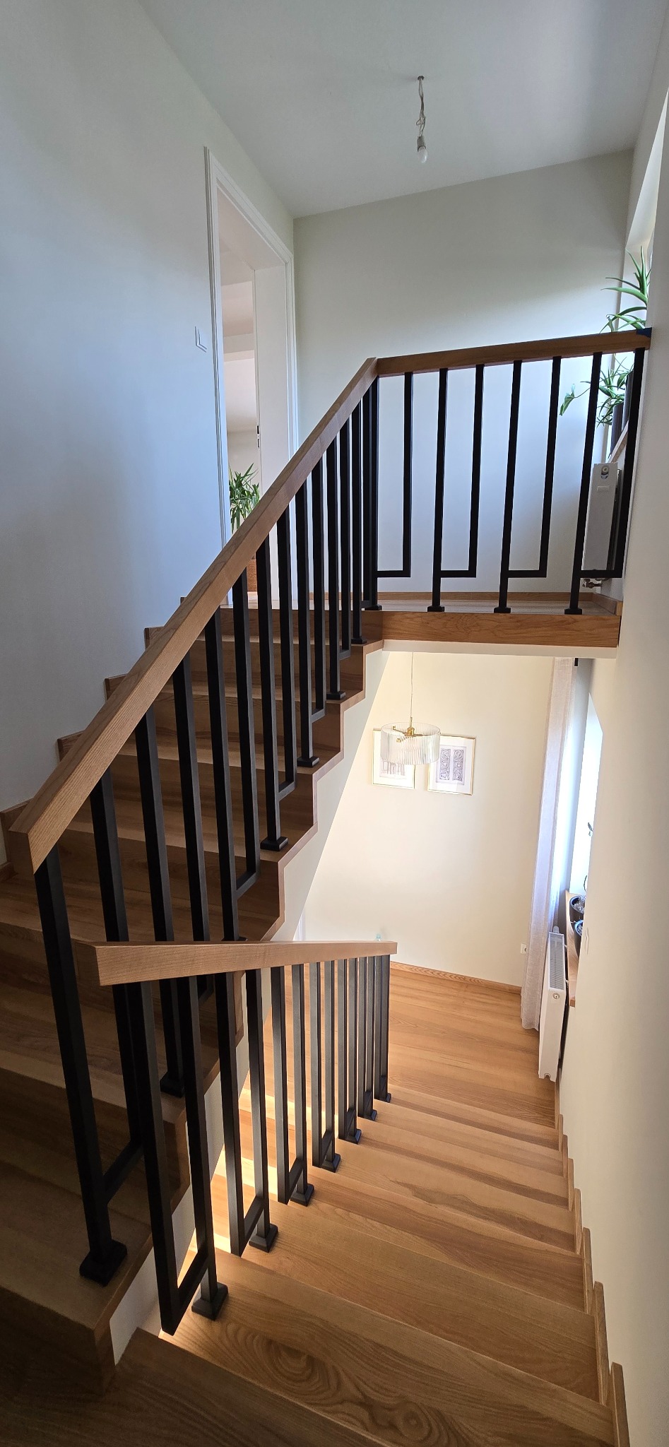 Jasne, drewniane schody z czarną, nowoczesną balustradą w jasnym wnętrzu. Widok z góry na dół, z delikatnym oświetleniem i rośliną w tle.