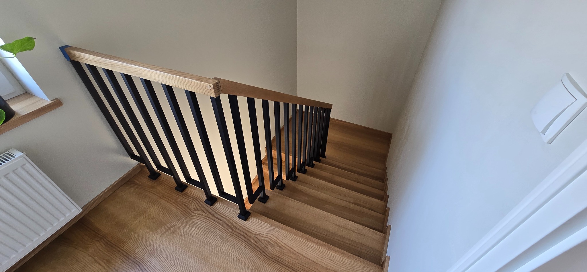 Nowoczesne schody z drewnianymi stopniami i czarną, metalową balustradą, widziane z góry. Jasne drewno kontrastuje z ciemnymi elementami metalowymi.