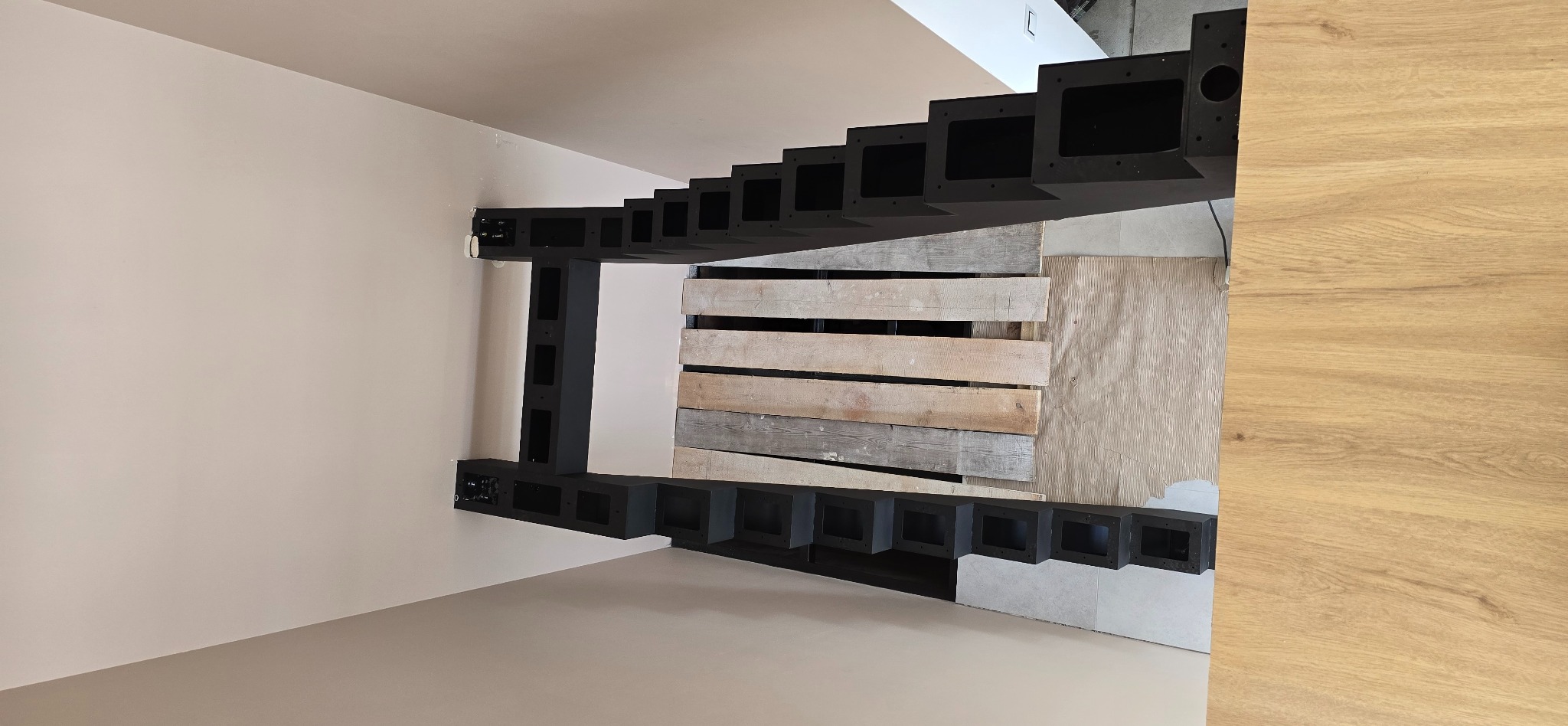 Częściowo zamontowana, czarna konstrukcja schodów z otworami, widok z dołu, surowe drewno w tle. Etap montażu, nietypowy kadr.