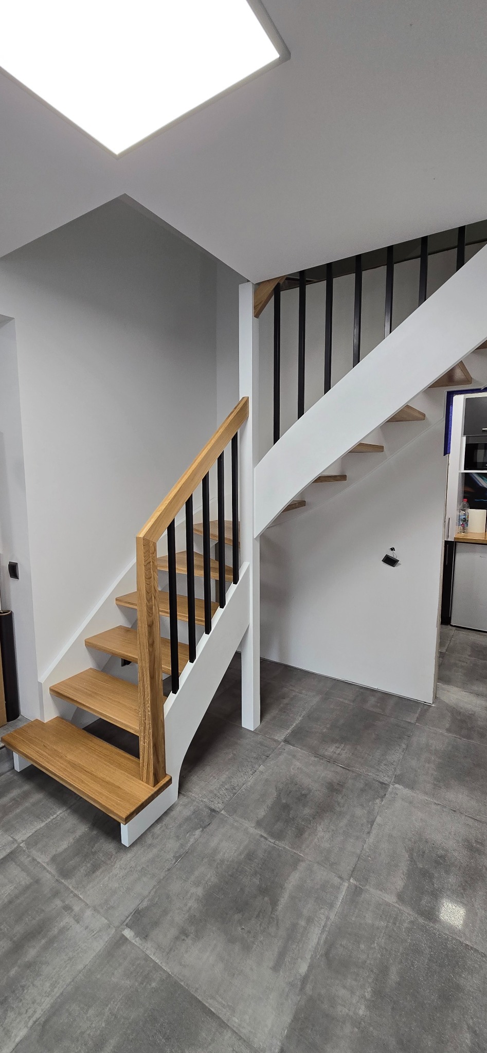 Nowoczesne schody drewniane z czarnymi balustradami i białymi elementami konstrukcyjnymi, na tle szarej podłogi z płytek. Widok z dołu, oświetlenie sufitowe.