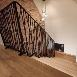 Nowoczesne schody drewniane z metalową, geometryczną balustradą na tle ściany z czerwonej cegły i minimalistycznych lamp wiszących.