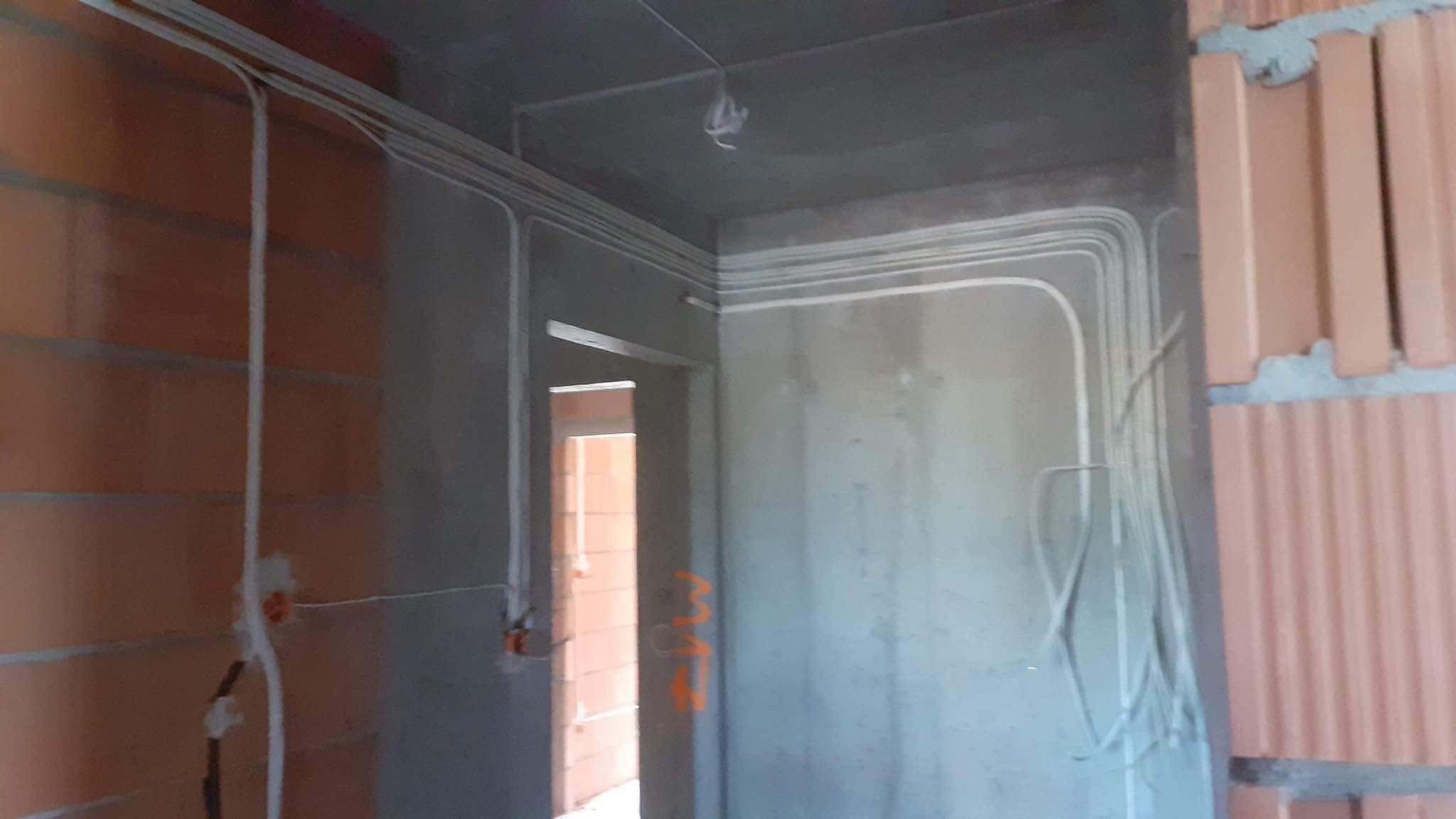 Surowe wnętrze w trakcie prac wykończeniowych z widocznymi przewodami elektrycznymi poprowadzonymi po ścianach i suficie, zamocowanymi w białych rurkach instalacyjnych.