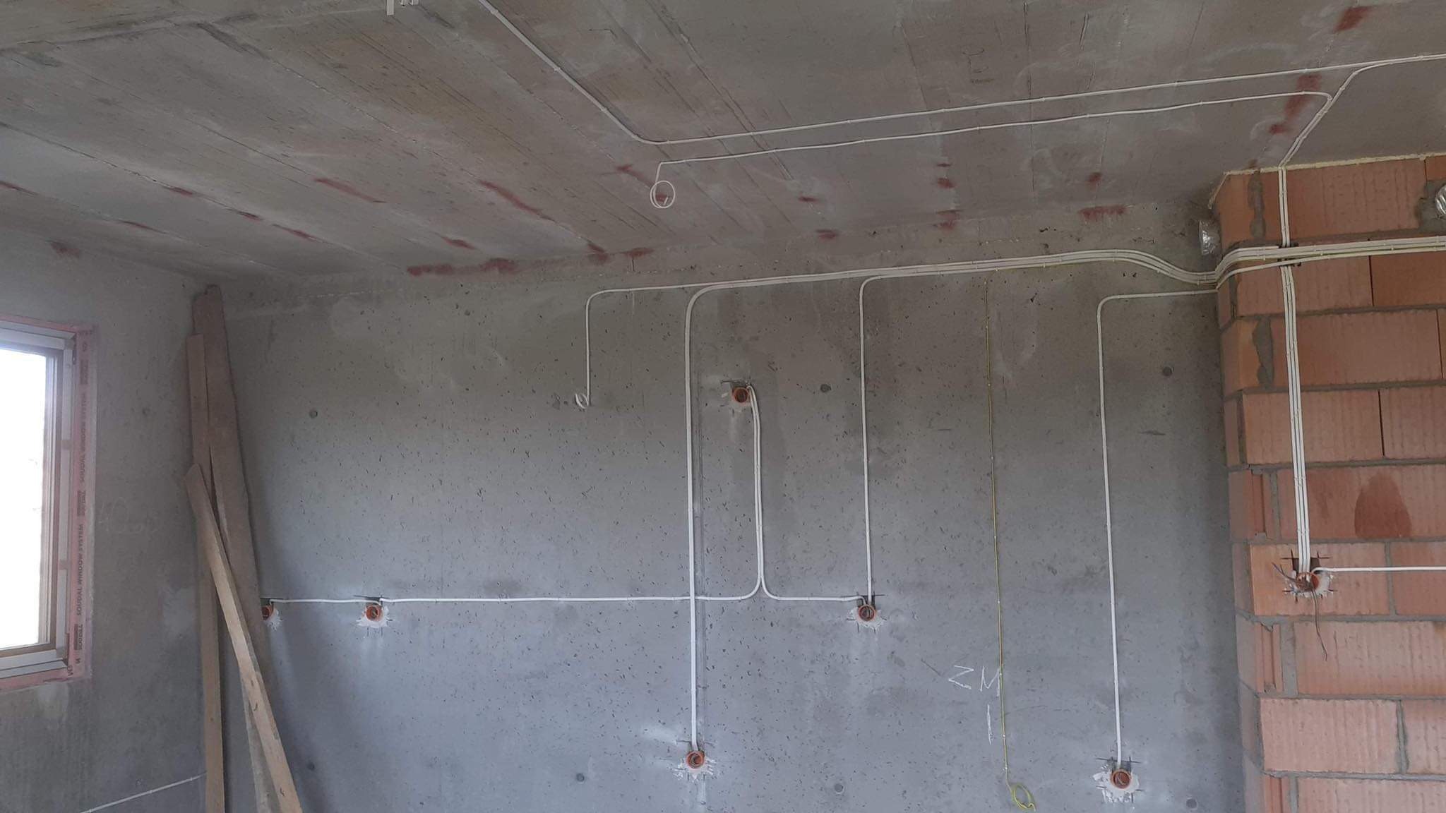 Surowy stan: Białe przewody elektryczne poprowadzone po szarym betonowym suficie i ścianach w nowo budowanym pomieszczeniu, widoczne puszki instalacyjne, fragment okna i ceglanego muru.