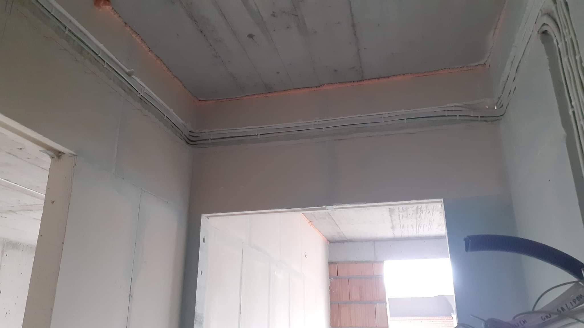 Montaż instalacji elektrycznej w surowym wnętrzu: kable prowadzone po ścianach i suficie, widoczne mocowania i fragment czarnej rury karbowanej.