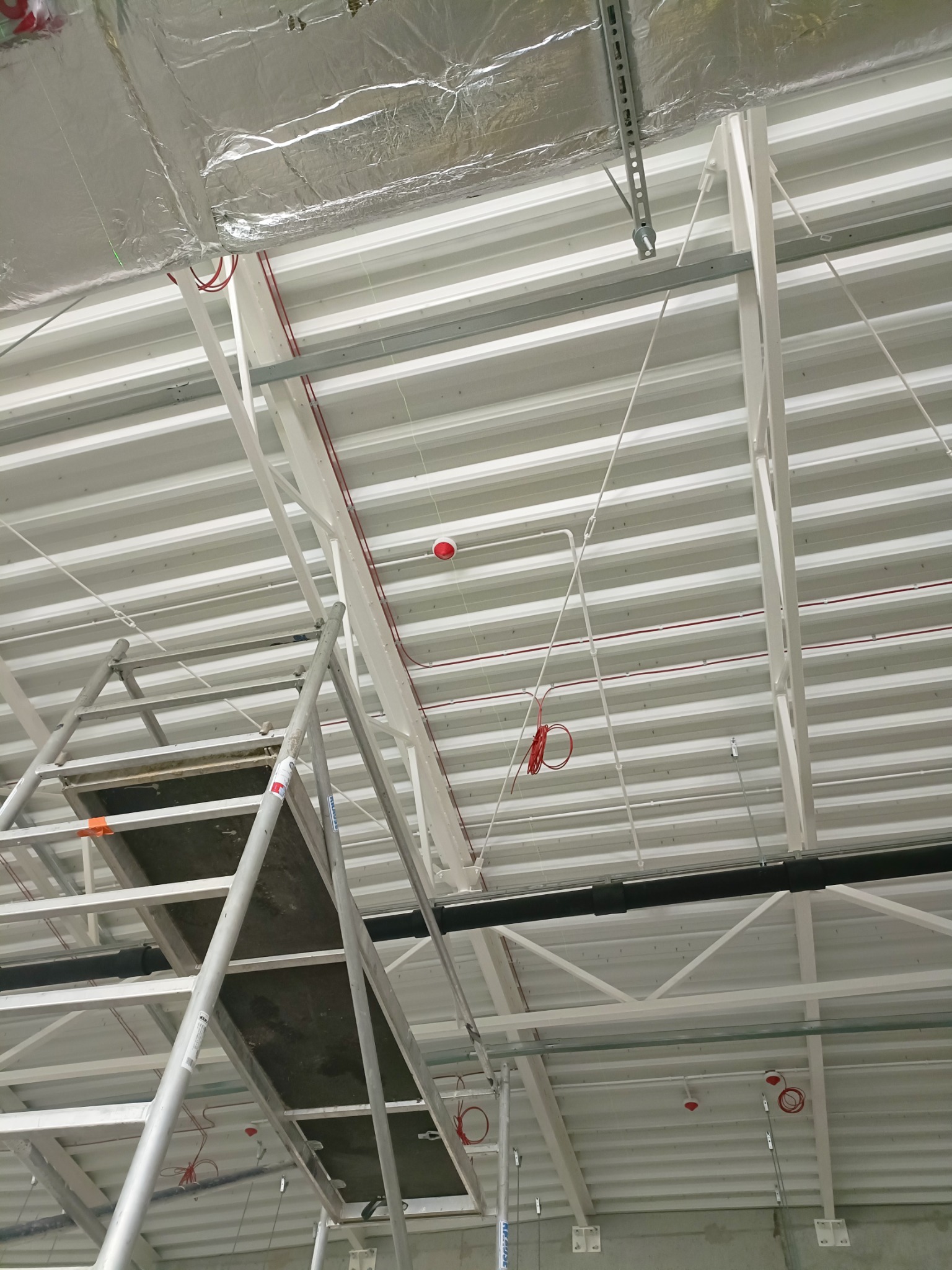 Sufit hali przemysłowej w trakcie instalacji elektrycznej, widoczne kable w peszlach, elementy oświetlenia awaryjnego, konstrukcja stalowa dachu i rusztowanie.