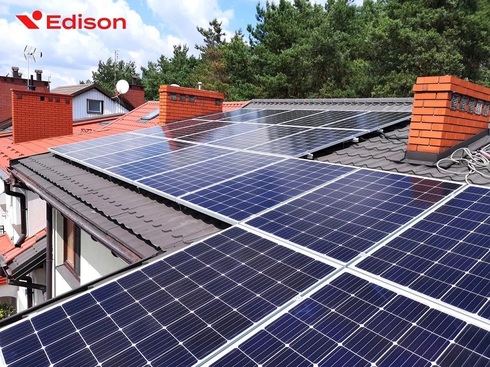 Panele słoneczne na ciemnym dachu domu jednorodzinnego z ceglanym kominem, widoczne logo firmy Edison w lewym górnym rogu.