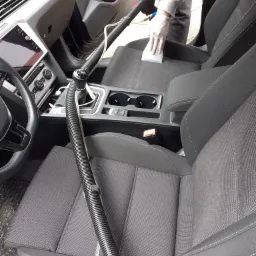 Czyszczenie tapicerki samochodowej: osoba w rękawicach czyści siedzisko szarą tapicerką przy użyciu odkurzacza z grubą rurą i białej ściereczki