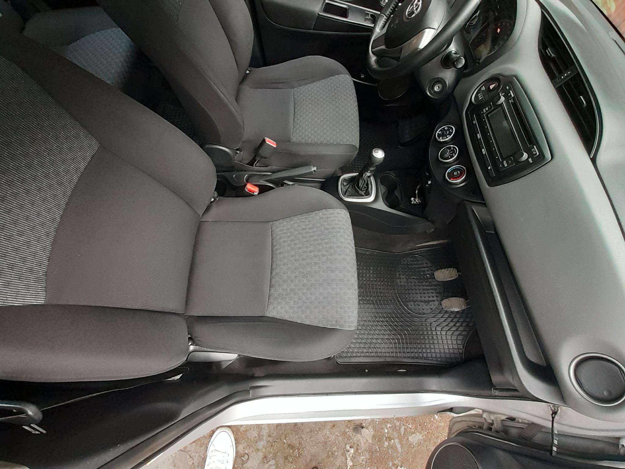 Wnętrze samochodu Toyota Yaris z widocznymi przednimi fotelami z szarej tapicerki, kierownicą i deską rozdzielczą. Dywaniki gumowe na podłodze. Widoczne fragmenty białego buta przy progu.