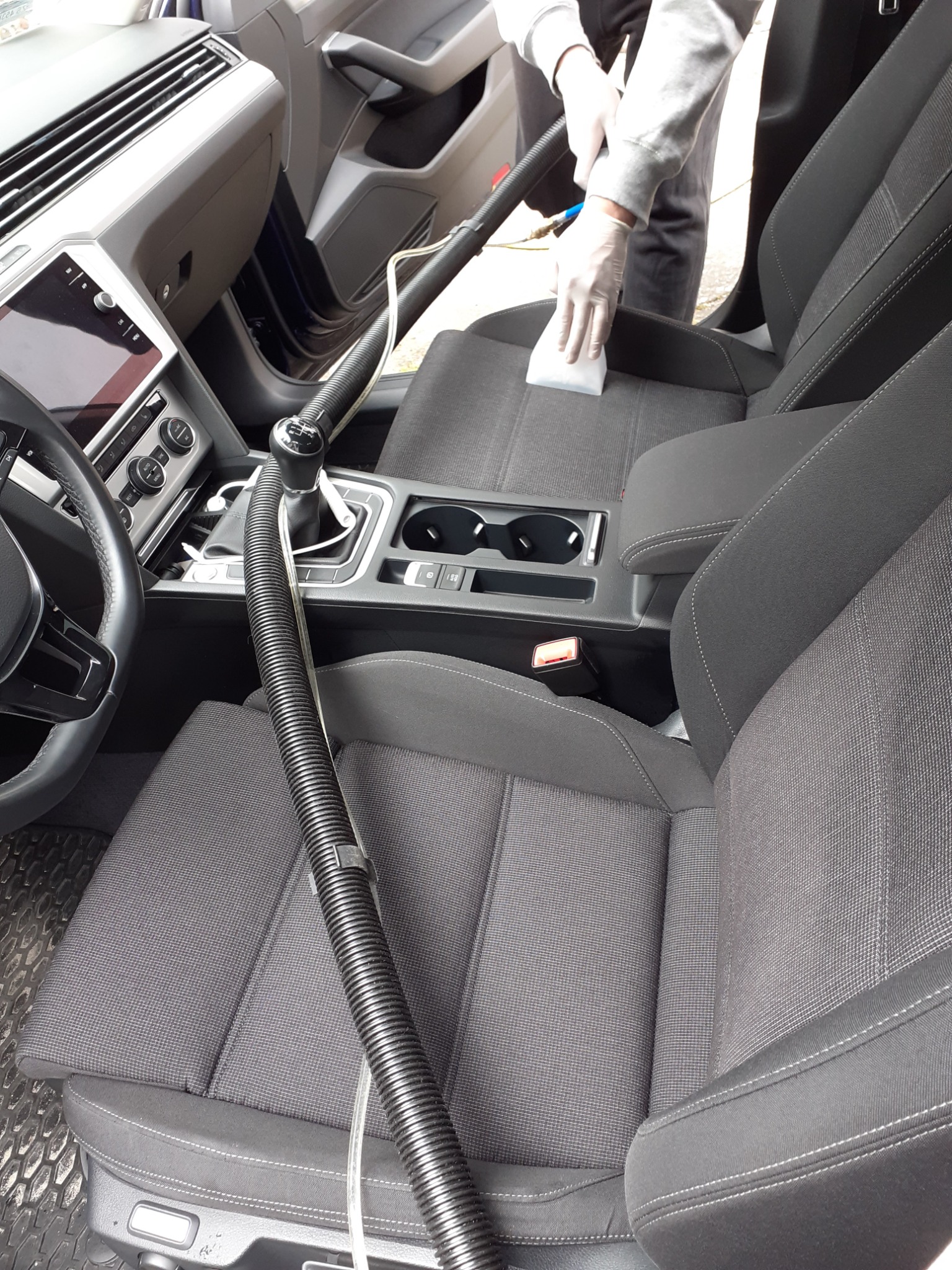 Czyszczenie tapicerki samochodowej: osoba w rękawicach czyści siedzisko szarą tapicerką przy użyciu odkurzacza z grubą rurą i białej ściereczki