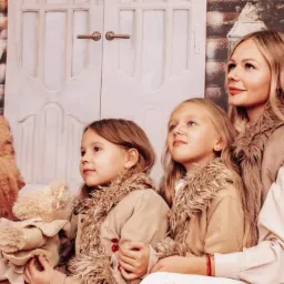 Portret matki z dwiema córkami w zimowej stylizacji na tle dekoracji świątecznej, dziewczynka trzyma pluszowego misia.