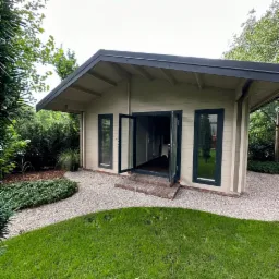 Nowoczesny, parterowy budynek z płaskim dachem i dużymi oknami, otoczony zielenią i żwirową ścieżką, z otwartymi dwuskrzydłowymi drzwiami wejściowymi i ceglanymi schodkami.