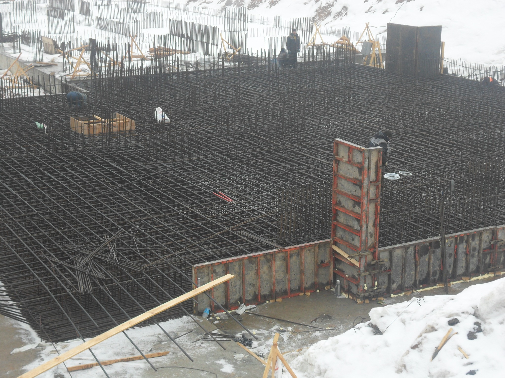 Zbrojenie fundamentu z widocznymi szalunkami i pracownikami w tle, teren budowy w zimowych warunkach.
