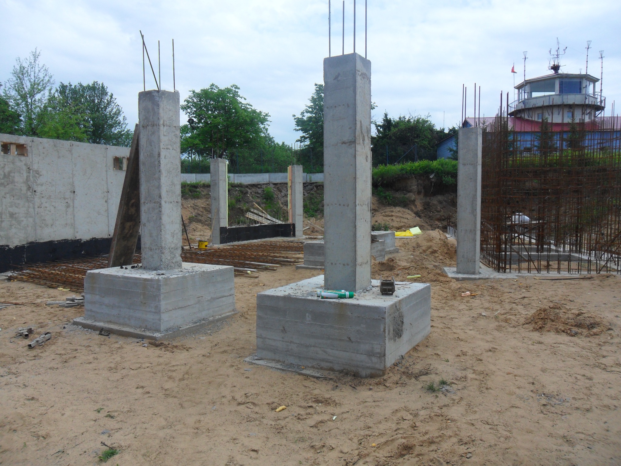Fundamenty kolumnowe z betonu z wystającymi prętami zbrojeniowymi na placu budowy, w tle budynek kontroli lotów.