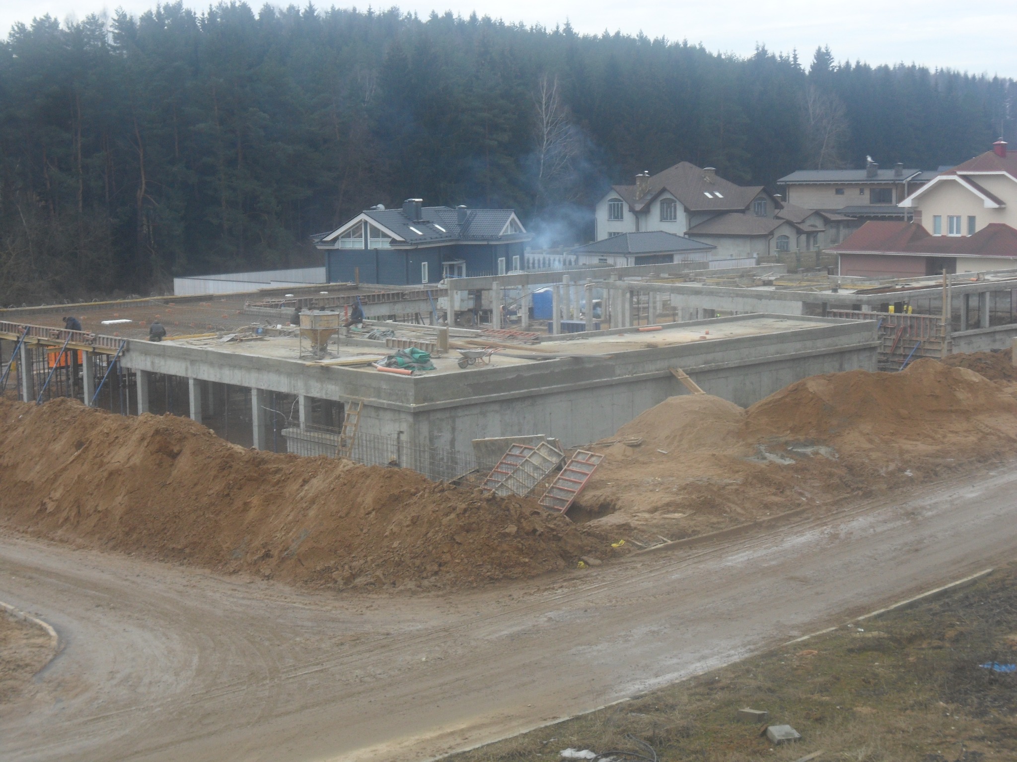 Stan surowy otwarty domu murowanego w trakcie budowy, widoczne fundamenty, ściany parteru, szalunki, hałdy ziemi i pobliskie domy jednorodzinne w tle.