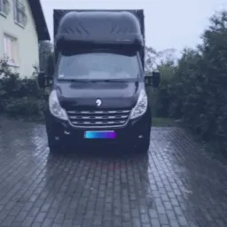 Czarny bus z wysoką zabudową stoi na brukowanym podjeździe przed domem w pochmurny dzień.