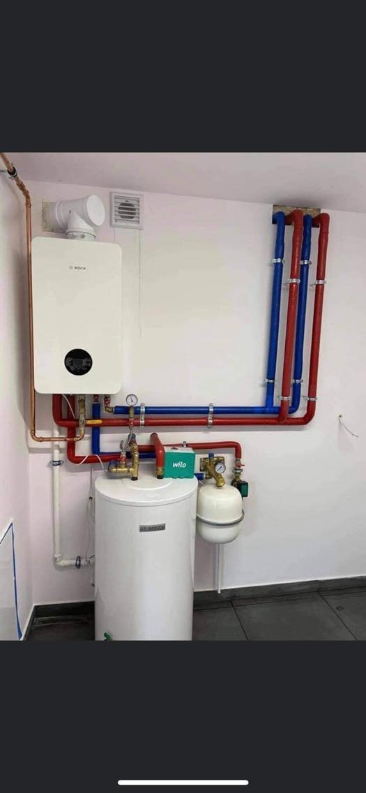 Biały piec gazowy Bosch z podłączonymi rurami miedzianymi i niebiesko-czerwonymi rurami instalacji grzewczej, biały zasobnik ciepłej wody z zielonym modułem Wito, szara płytka podłogowa.