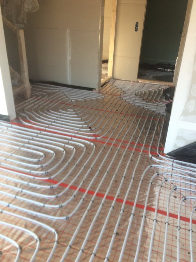 Białe rurki ogrzewania podłogowego ułożone w pętle na folii z czerwoną siatką, widoczne ściany w surowym stanie.