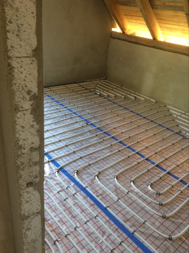 Białe rurki ogrzewania podłogowego ułożone na folii z czerwoną siatką, przymocowane niebieską taśmą do podłoża w surowym stanie, widok z perspektywy wejścia.