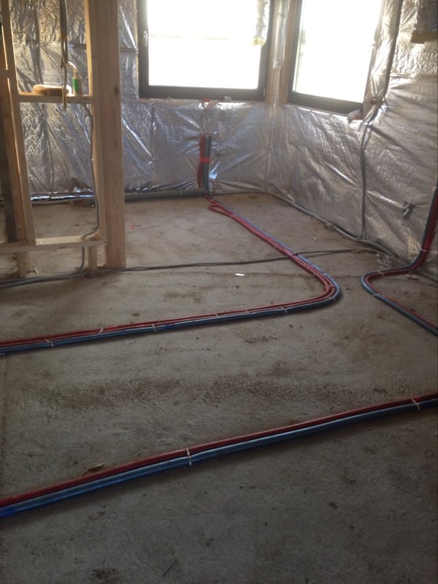 Surowe wnętrze w trakcie budowy z widocznymi rurami grzewczymi ułożonymi na posadzce, ściany pokryte folią izolacyjną.