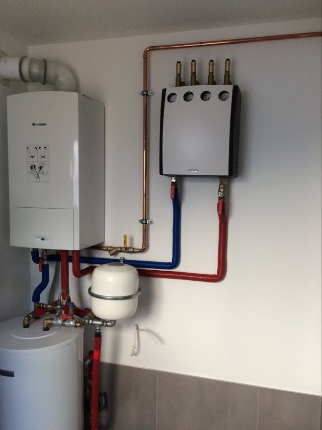 Zestaw urządzeń grzewczych zamontowany na ścianie: biały piec dwufunkcyjny, naczynie przeponowe i rozdzielacz z mosiężnymi zaworami, połączone rurami miedzianymi i z tworzywa w kolorach czerwonym...