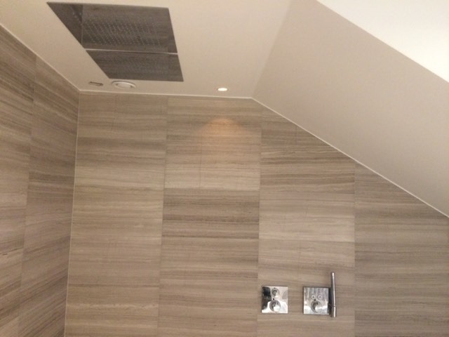 Nowoczesna kabina prysznicowa z deszczownicą w suficie, wykończona płytkami imitującymi drewno, z widocznymi bateriami podtynkowymi.