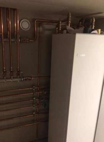 Instalacja centralnego ogrzewania z miedzianymi rurami i białym piecem, widoczne zawory i okrągły odpowietrznik na szarej ścianie.