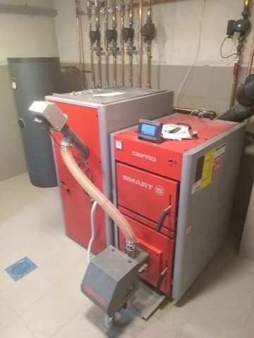 Czerwono-szary piec grzewczy Defro Smart z podajnikiem, zbiornikiem na wodę i instalacją rurową w kotłowni.