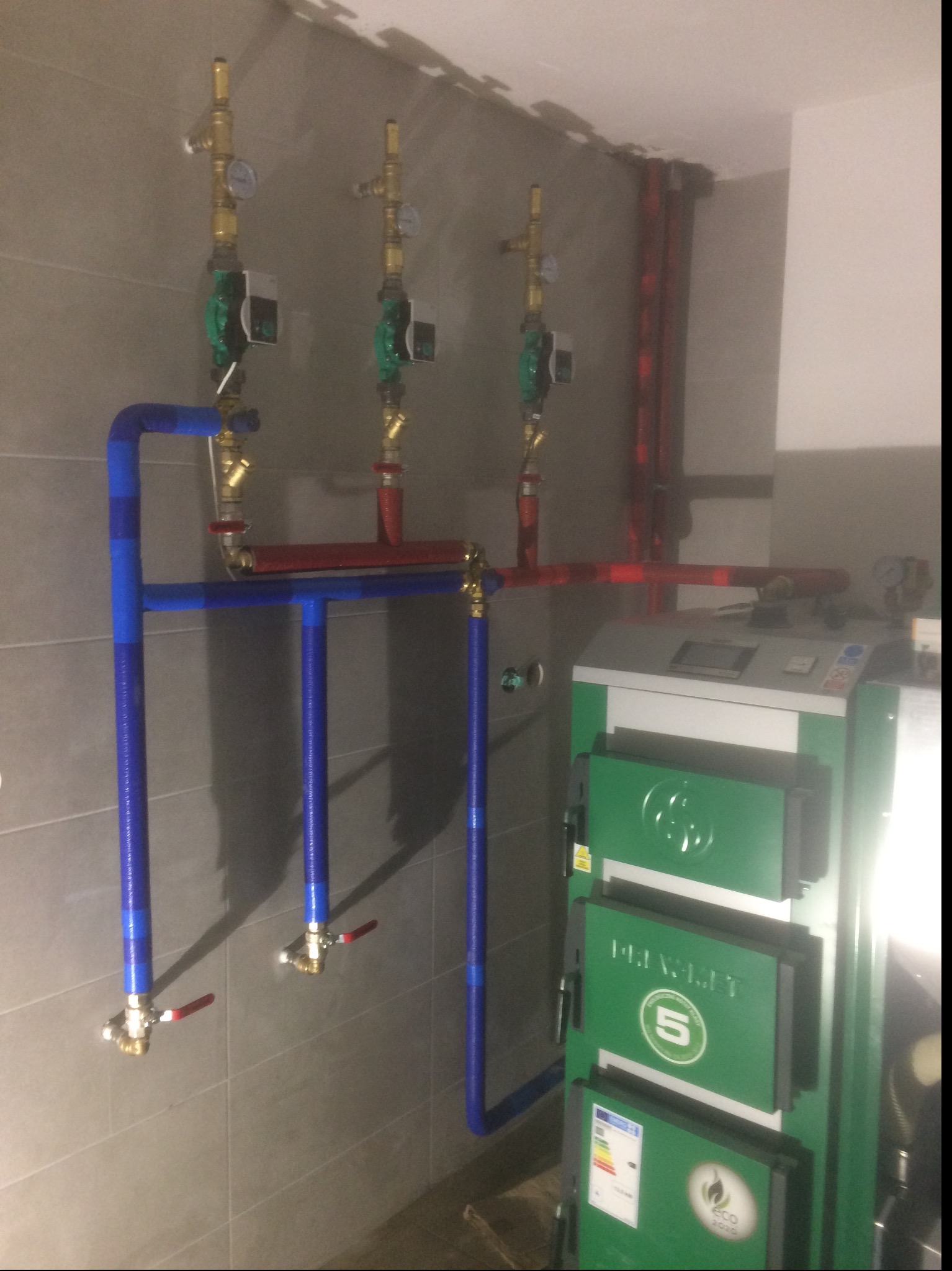 Instalacja kotła grzewczego z rurami w kolorach niebieskim i czerwonym, z zaworami i manometrami, na tle szarej ściany z płytek.