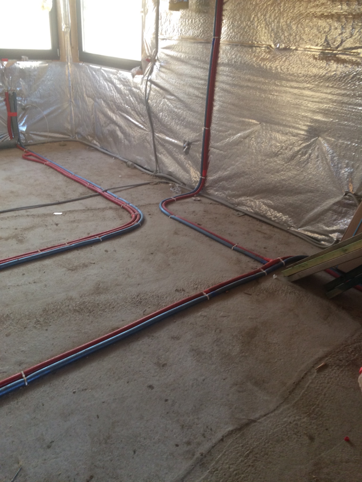 Rury wodne i kanalizacyjne prowadzone po podłodze w pomieszczeniu z izolacją termiczną na ścianach, zabezpieczone białymi opaskami zaciskowymi.