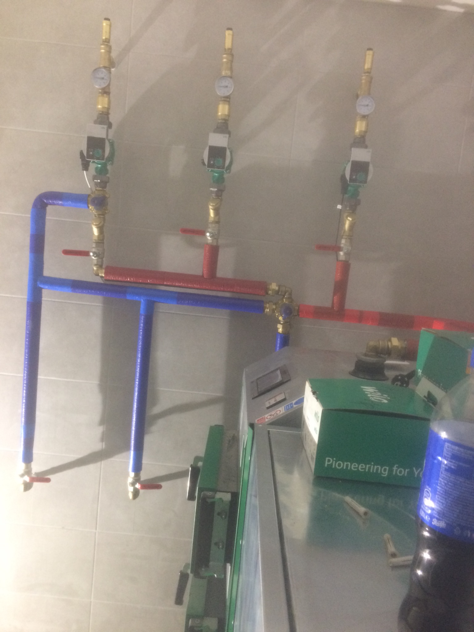 Trzy pionowe rury z manometrami i pompami obiegowymi, połączone poziomymi rurami w kolorach niebieskim i czerwonym, widoczne pudełka i butelka.