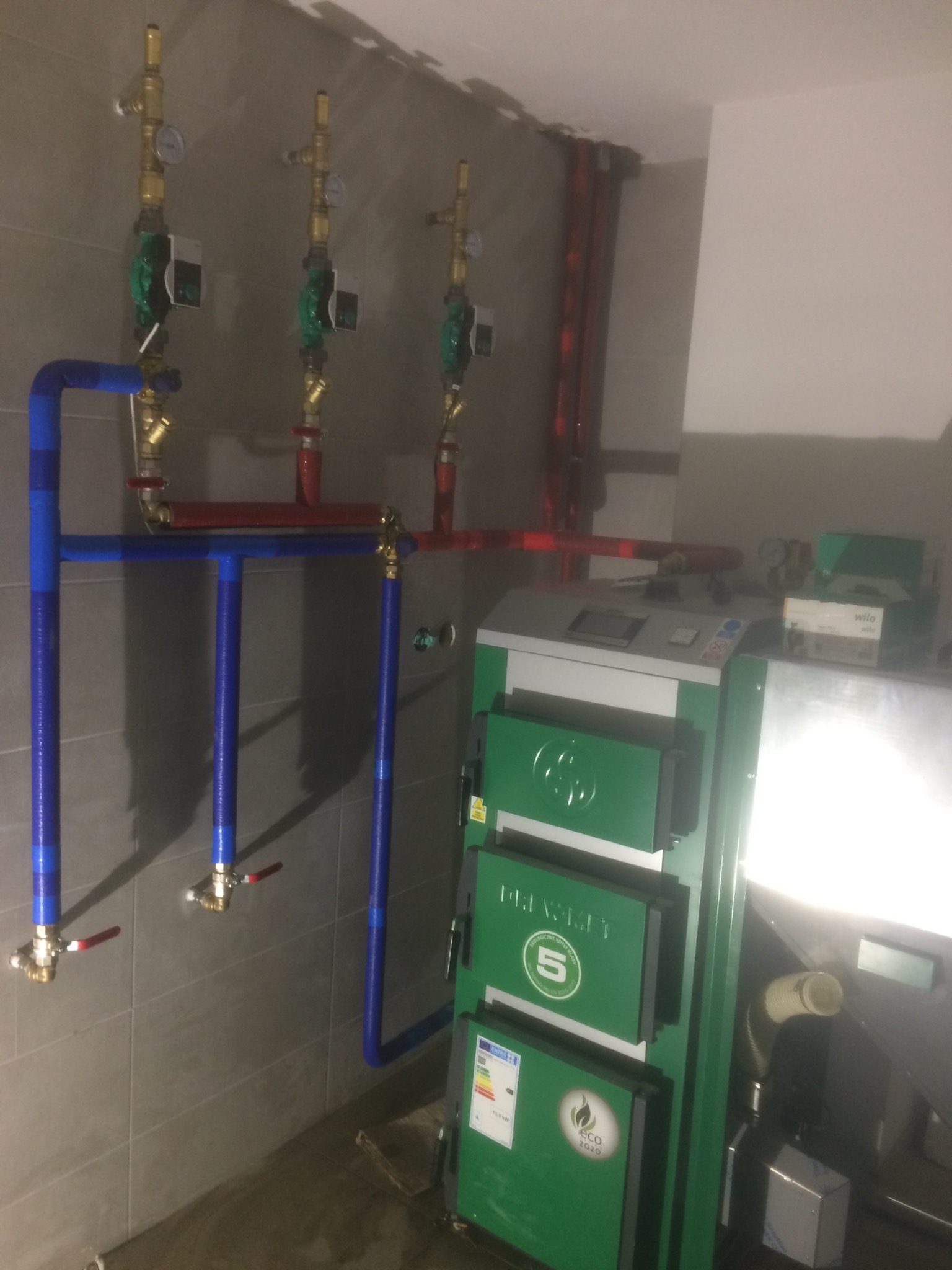 Instalacja centralnego ogrzewania z kotłem na paliwo stałe, widoczne rury w izolacji koloru niebieskiego i czerwonego, zawory oraz kocioł marki Prawet z oznaczeniem Eco 2020.