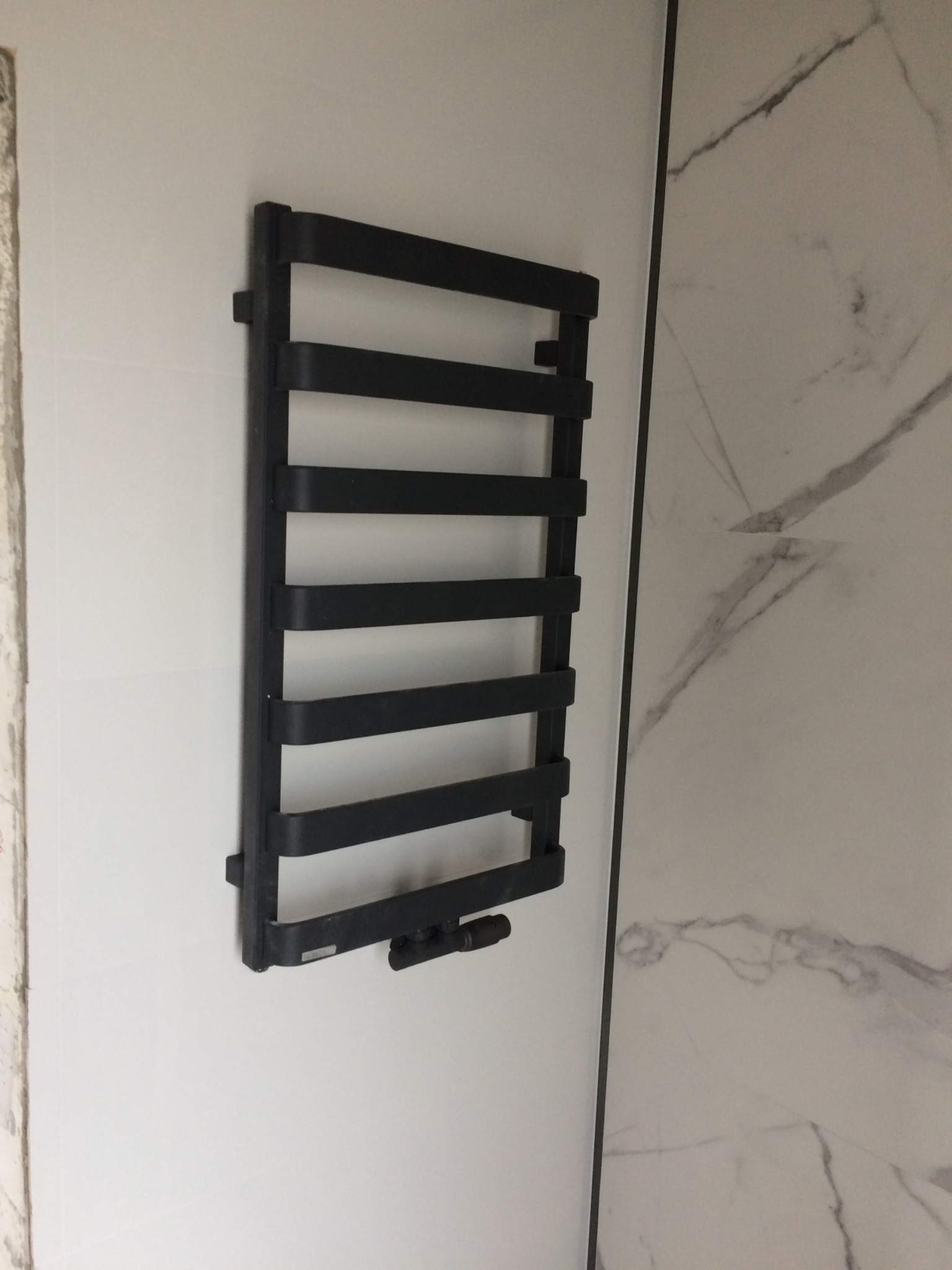 Czarny, drabinkowy grzejnik łazienkowy zamontowany na białej ścianie, obok ściany wykończonej płytkami imitującymi marmur.