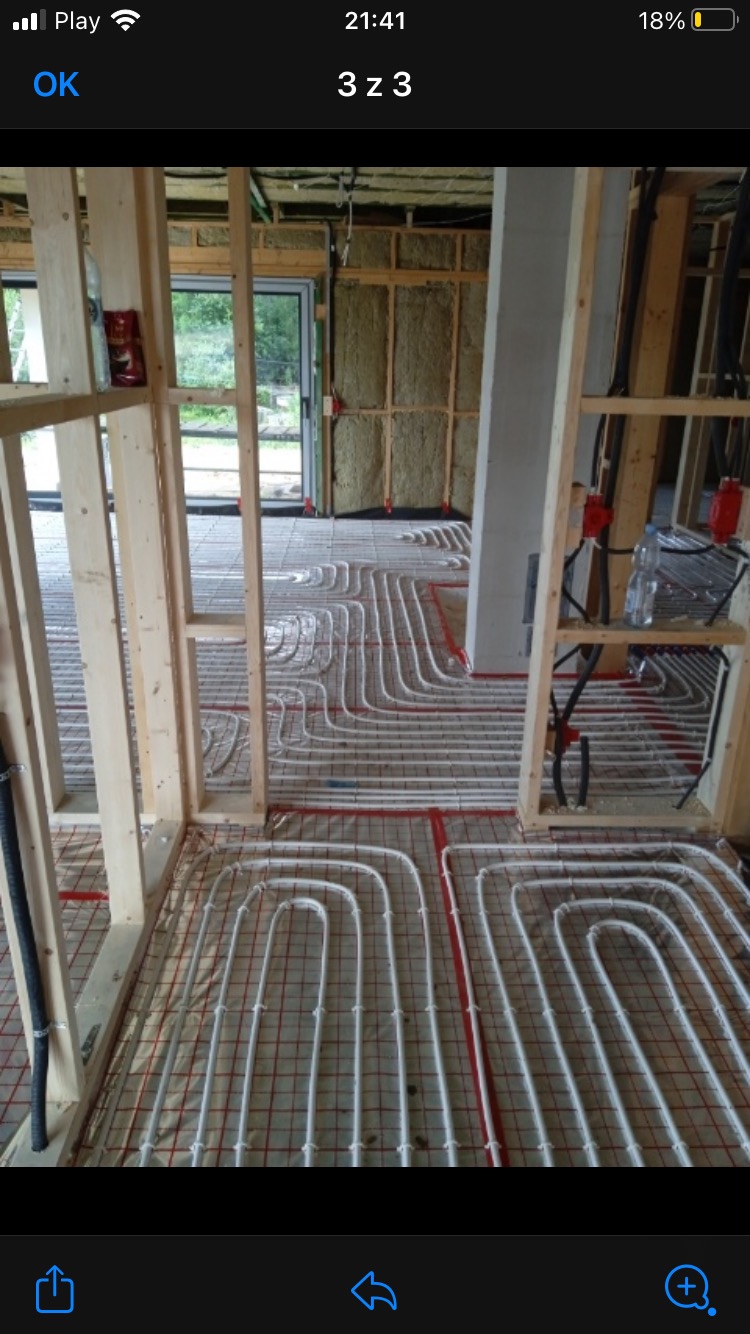 Białe rury ogrzewania podłogowego ułożone na siatce w surowym wnętrzu budynku w trakcie budowy, widoczne drewniane ramy konstrukcyjne i izolacja ścian wełną mineralną.