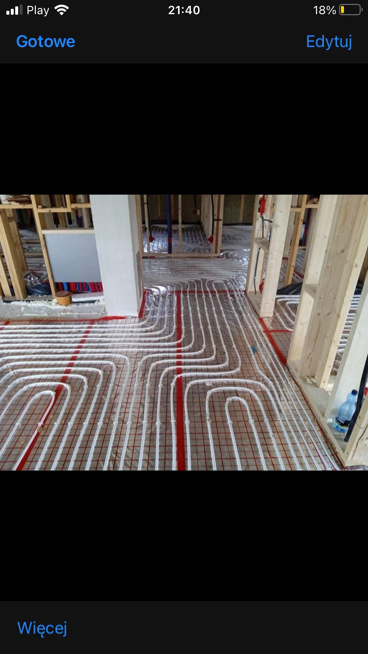 Instalacja ogrzewania podłogowego w trakcie montażu, rury ułożone na folii z czerwoną siatką, widoczne drewniane elementy konstrukcyjne budynku, butelka wody w rogu pomieszczenia.