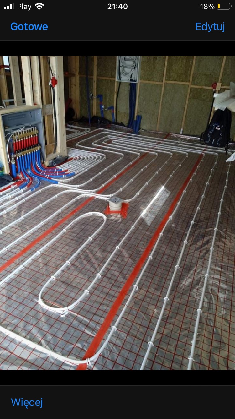 Rozłożone białe rury ogrzewania podłogowego na siatce z czerwonymi paskami, widoczny rozdzielacz z niebieskimi i czerwonymi złączkami, ściany w surowym stanie z widoczną izolacją.