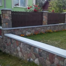 Ogrodzenie z brązowych desek z kamiennymi słupkami i podmurówką z kamienia polnego, otoczone zielenią i kwiatami, widok z niskiej perspektywy.