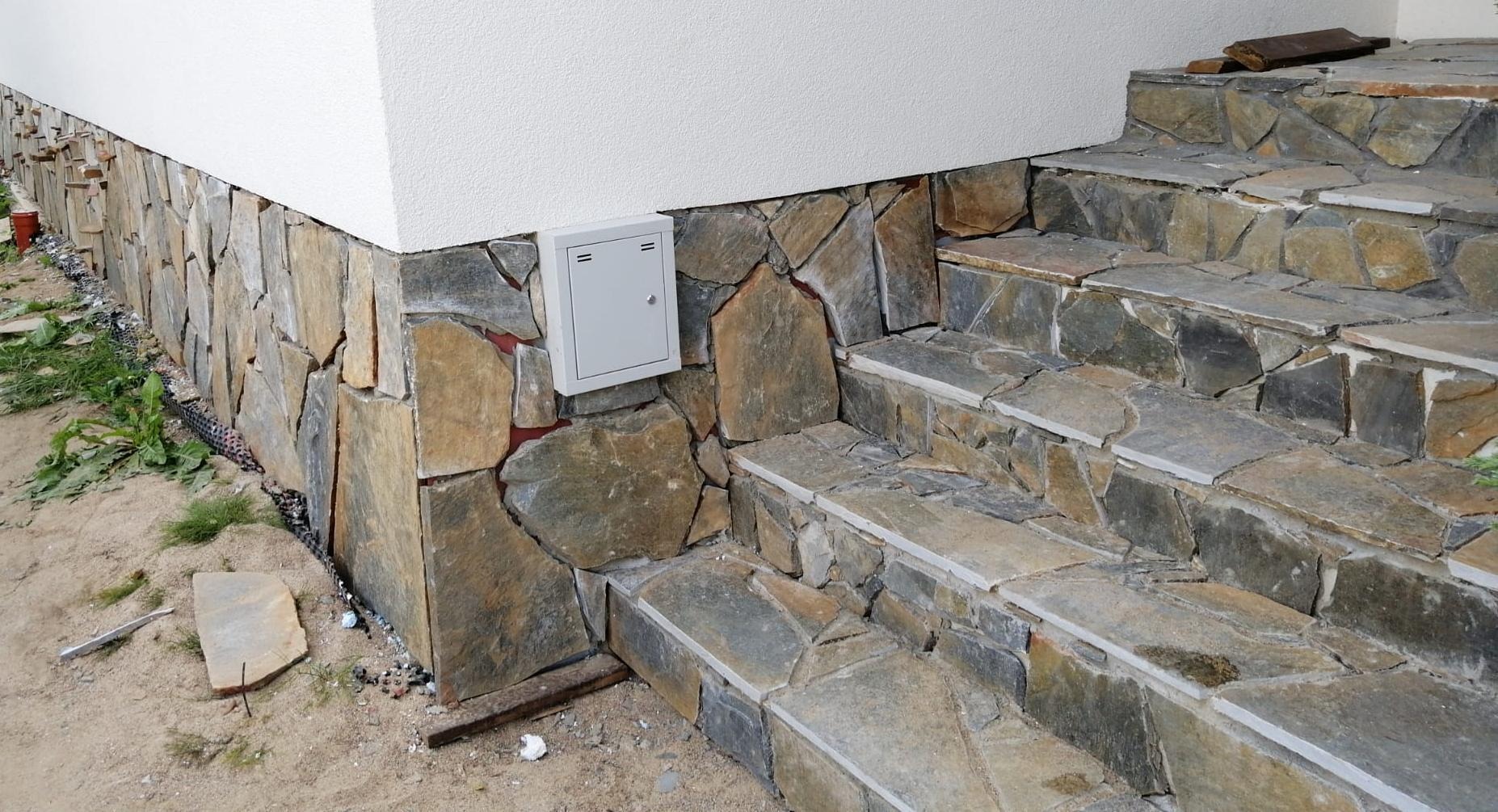 Kamienne schody wejściowe do domu, z okładziną z nieregularnych płyt kamiennych na cokole budynku i stopniach, obok biała skrzynka instalacyjna na elewacji.