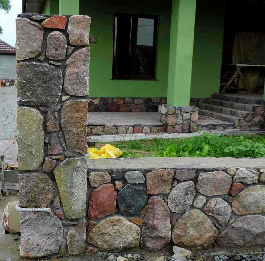 Kamienny taras z filarem z nieregularnych kamieni w odcieniach szarości, brązu i czerwieni, widok na wejście do domu z zielonymi kolumnami i schodami.