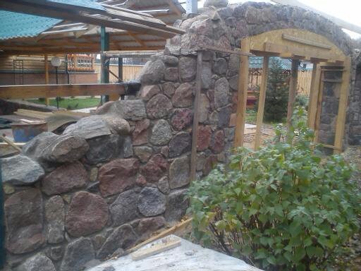 Fragment tarasu w trakcie budowy z elementami kamiennymi i drewnianą konstrukcją z widocznym zarysem łuku, otoczony zielenią.