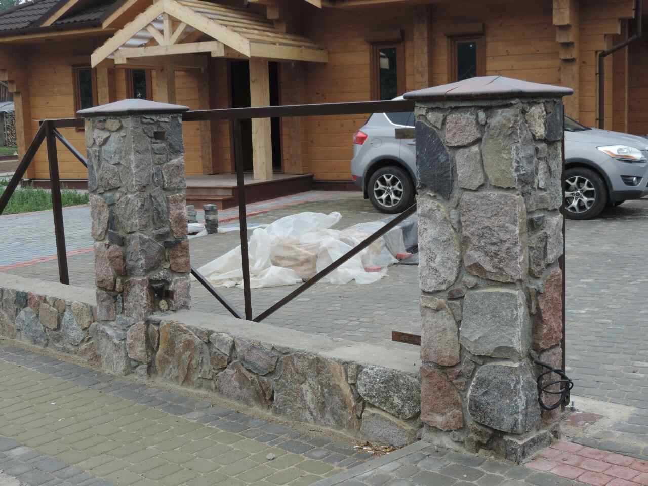 Fragment ogrodzenia z kamiennymi słupkami i metalowymi przęsłami, w tle drewniany dom i zaparkowany samochód, widoczne materiały budowlane zabezpieczone folią.