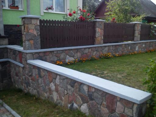 Ogrodzenie z brązowych desek z kamiennymi słupkami i podmurówką z kamienia polnego, otoczone zielenią i kwiatami, widok z niskiej perspektywy.