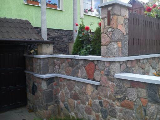 Kamienny murek ogrodzeniowy z filarami, z szarymi elementami wykończeniowymi, widok z boku, w tle zielony dom i drewniane ogrodzenie.