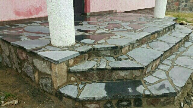 Kamienne schody wejściowe do domu, wykonane z nieregularnych płyt kamiennych w odcieniach szarości, z widoczną teksturą kamienia i fug, obok kolumny werandy.