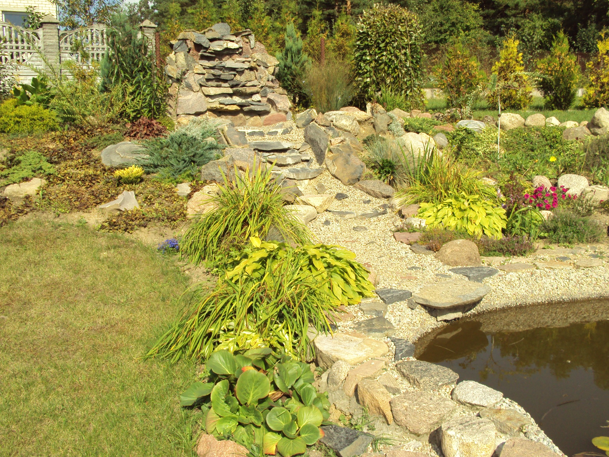Ogród z kaskadą wodną zbudowaną z płaskich kamieni, otoczoną roślinnością, trawą i małym stawem z kamiennym brzegiem.