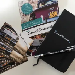 Zestaw materiałów reklamowych firmy oferującej wynajem samochodów do ślubu: wizytówki z danymi kontaktowymi, folder z ofertą i czarny notatnik z długopisem, wszystko na białym tle.