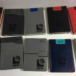 Kolekcja dziesięciu notesów firmowych z logo NGK i rokiem 2019, w różnych kolorach okładek: czerwonym, szarym, czarnym i niebieskim, ułożonych na białym tle.